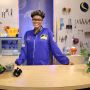 Netia McCray is shown wearing a blue jumpsuit and standing behind a table that has several plants on it. Behind her is a peg board wall with tools.